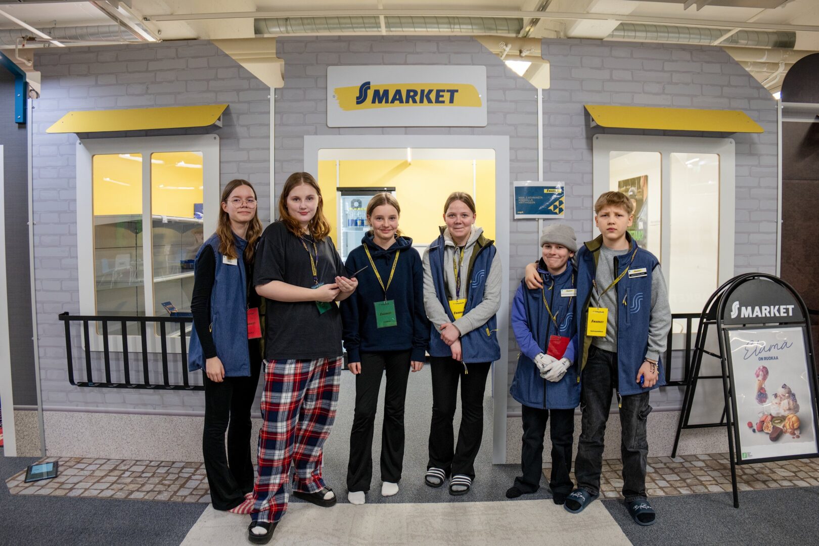 Viisi nuorta seisoo rinnakkain S-Marketin edessä Yrityskylässä ja katsoo kameraan.