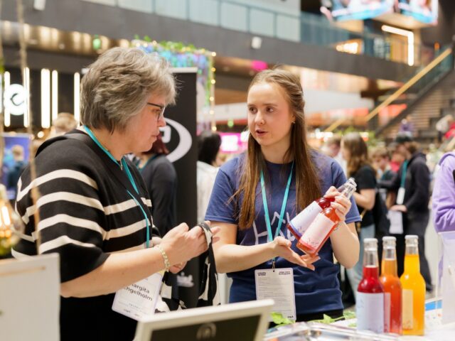 Nuori henkilö esittelee NYT-yrityksensä tuotteita, kahta virvoitusjuomapulloa, asiakkaalle.