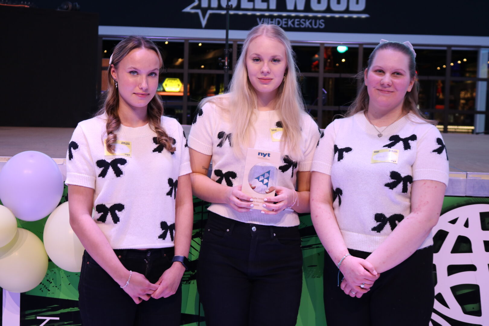 Kolme nuorta samanlaisissa mustavalkoisissa paidoissa seisoo rivissä, keskimmäinen pitää käsissään pokaalia.