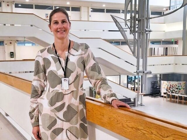 Tiina Mätäsjärvi nojaa kaiteeseen, taustalla koulun portaikkoa ja aulatilaa.