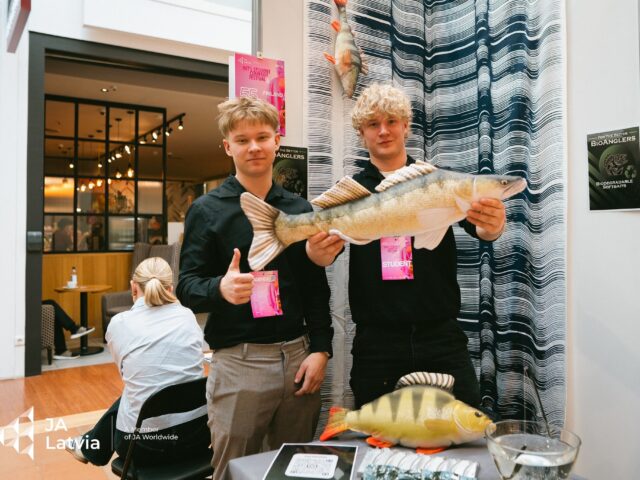 Kaksi nuorta seisoo messupisteellä ja esittelee suurta muovista kalaa kameralle.