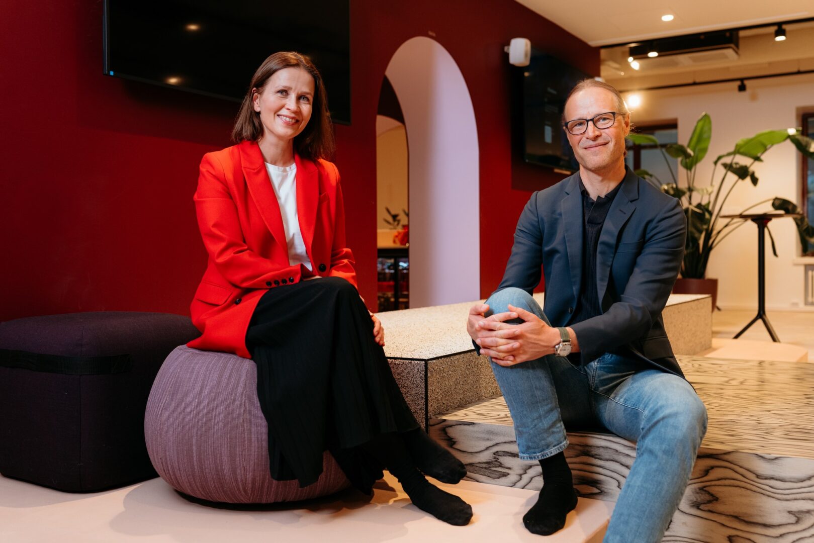 Kaksi henkilöä katsoo kameraan, istuvat korokkeilla lounge-tilassa.