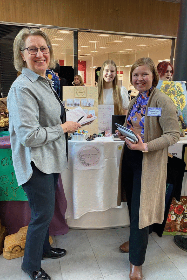 Kaksi naishenkilöä seisoo nuoren yrittäjän messupisteen edessä ja hymyilee kameralle.