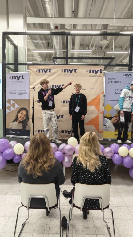 Kaksi nuorta seisoo lavalla ja pitää myyntipuhetta tuomareille, jotka kuuntelevat selin kameraan.