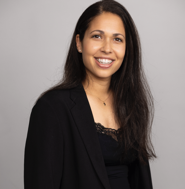 Jakkupukuun pukeutunut nuori henkilö katsoo kameraan ja hymyilee.