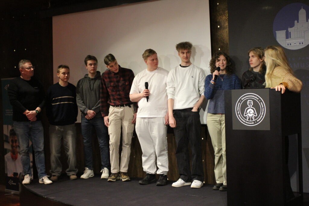 Ryhmä nuoria seisoo lavalla rivissä ja kahdella on käsissään mikrofoni.