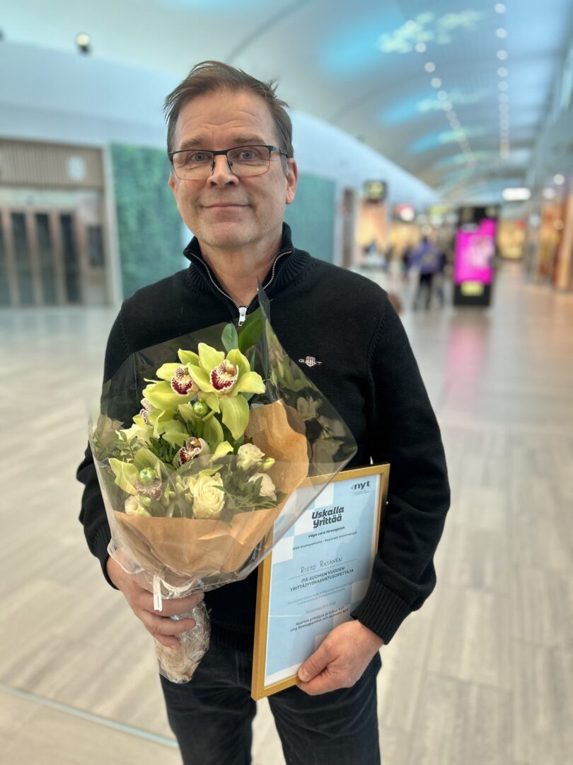 Henkilö hymyilee kameralle ja pitää kädessään kukkia sekä palkintoplakaatia.
