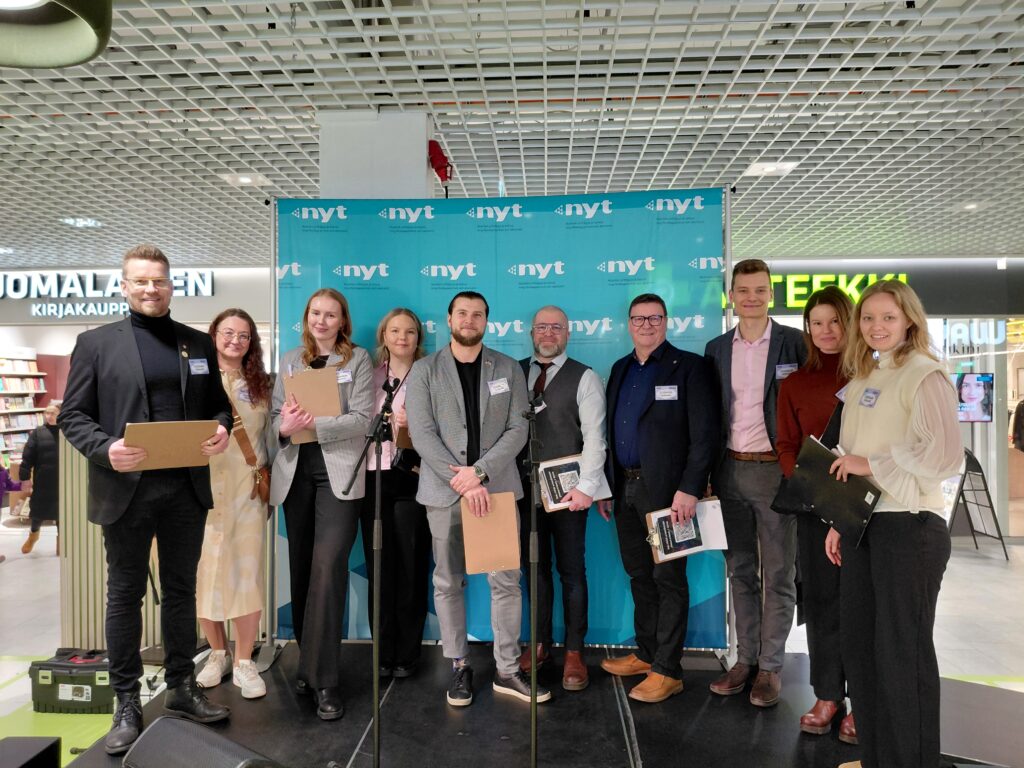 Joukko henkilöitä seisoo rivissä lavalla katsoen kameraan.