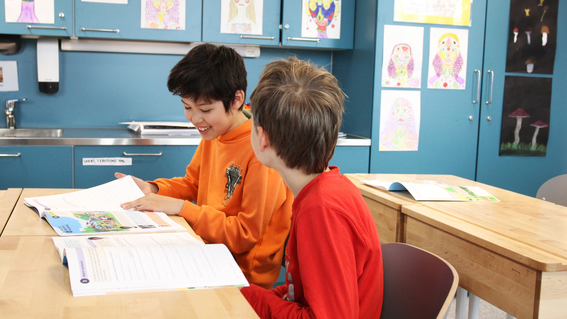 Kaksi lasta istuu pulpettien ääressä ja selaa tehtäväkirjoja.