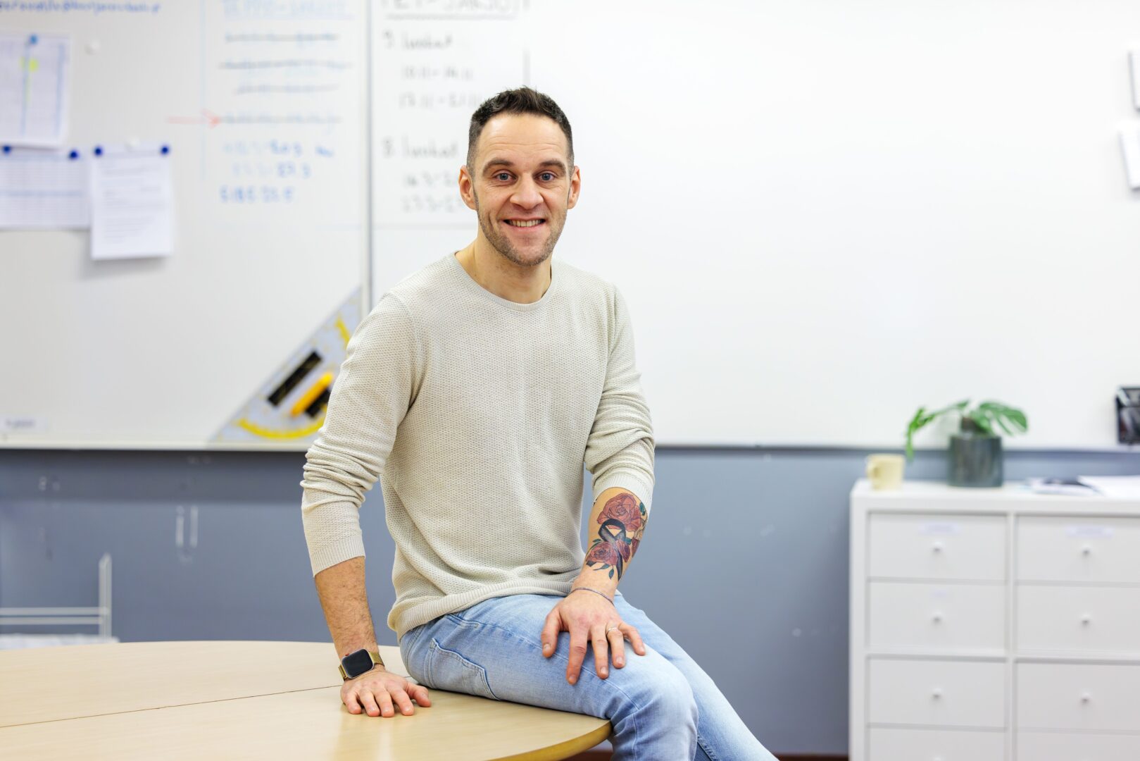 Henkilö istuu pöydän kulmalla opettajanhuoneessa ja katsoo kohti kameraa.