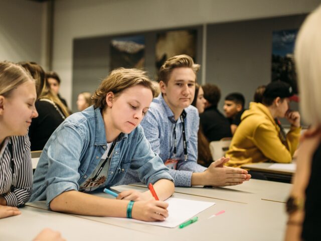 Kolme nuorta pöydän äärellä. Keskimmäinen nuorista kirjoittaa muistiinpanoja